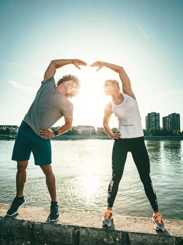 young-happy-couple-is-training-outdoors-by-river-stretching-together-wall-sunset